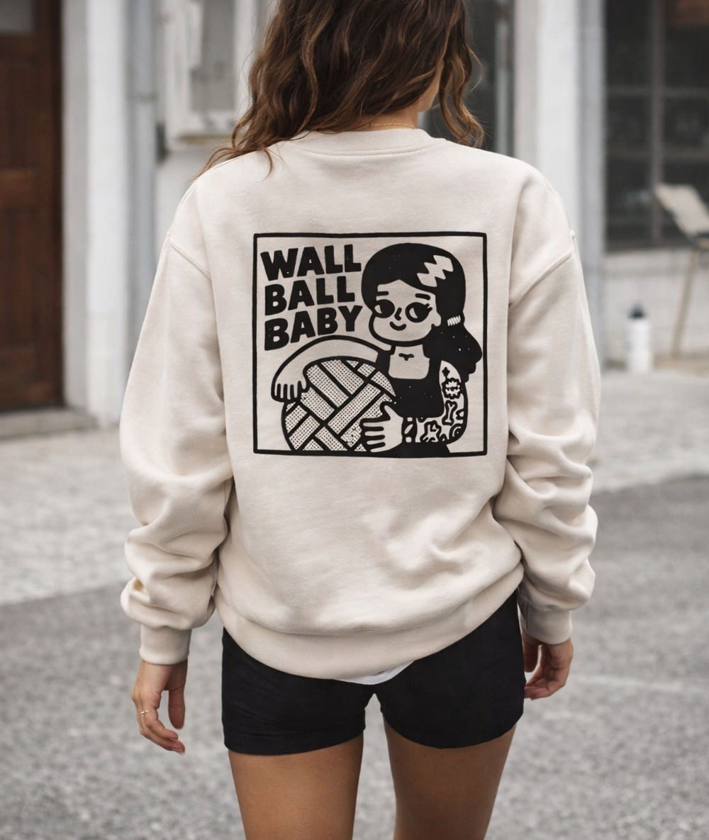 Person wearing a beige sweatshirt with 'Wall Ball Baby' graphic design outdoors.